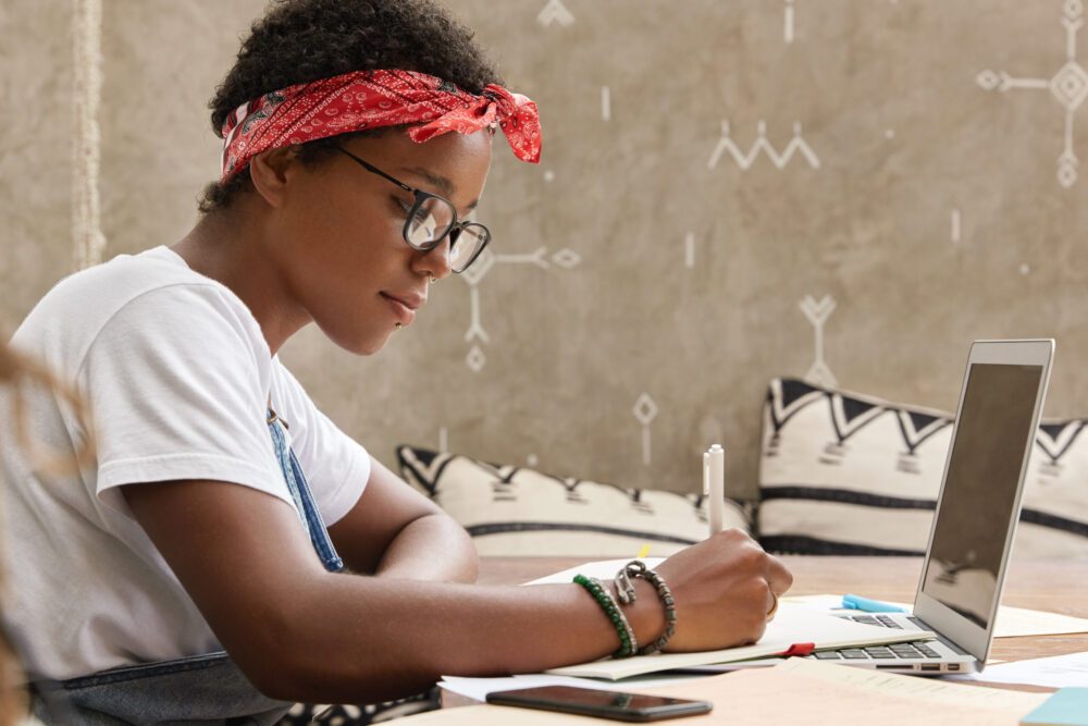 shot of serious african american student makes notes for making research, prepares report on particular topic, dressed in casual clothes, sits in front of laptop, learns information from internet